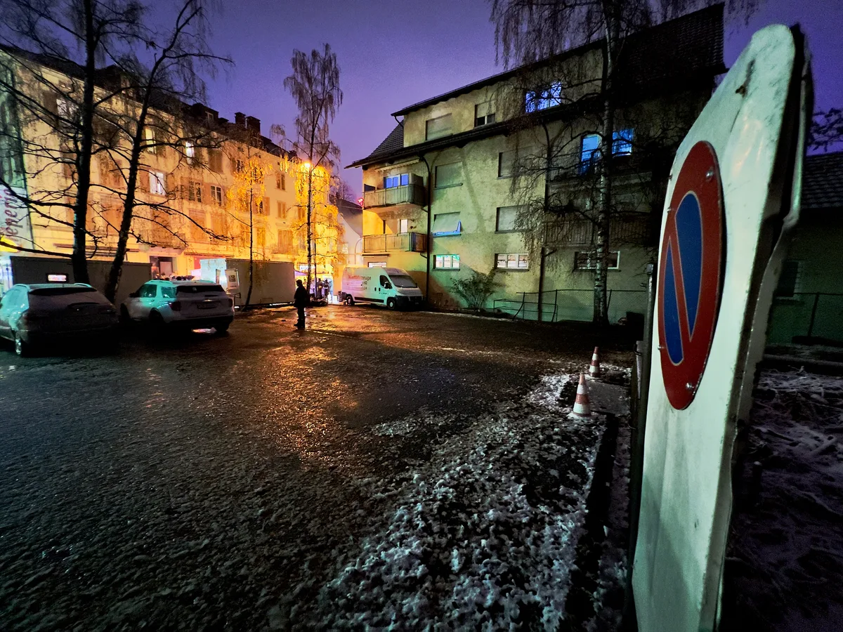 Man sieht einen leeren Platz und ein Parkverbotsschild. Ein paar Autos stehen auf der gegenüberliegenden Seite. Im Hintergrund sind städtische Gebäude zu sehen.